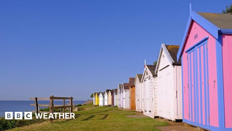 A row of multi-coloured beach huts on the right with some grass and a view over looking blue sea and bright blue sky